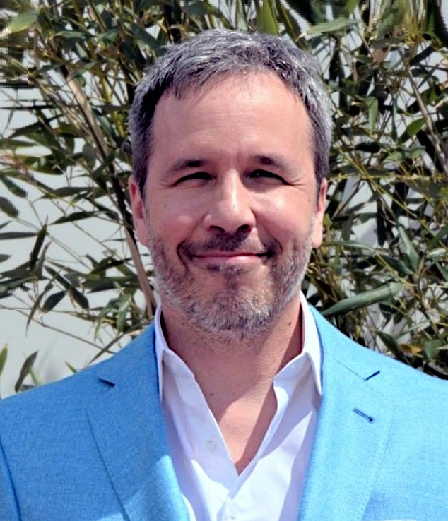 Portrait photo of Denis Villeneuve smiling outdoors in a light blue suit jacket and white shirt.