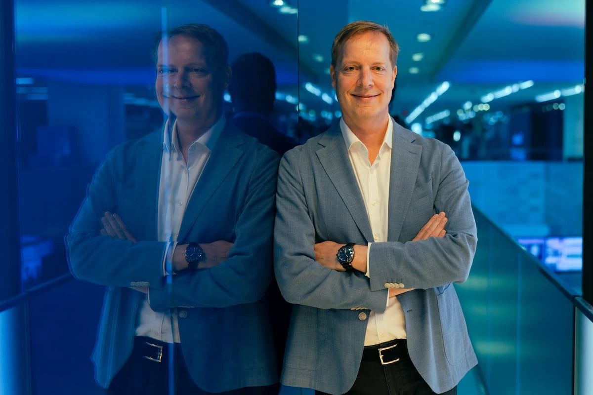 Portrait of Almar Latour in a blue suit, standing with arms crossed in a modern office setting.