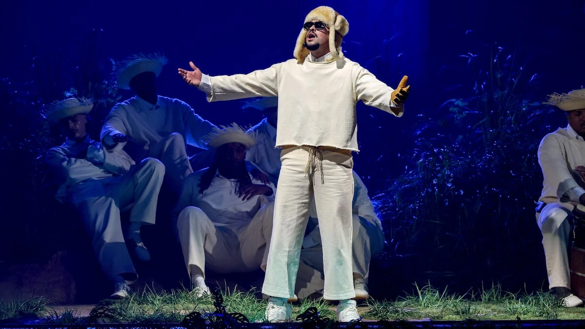 Bad Bunny performing on stage in a white outfit with arms outstretched under blue lighting.