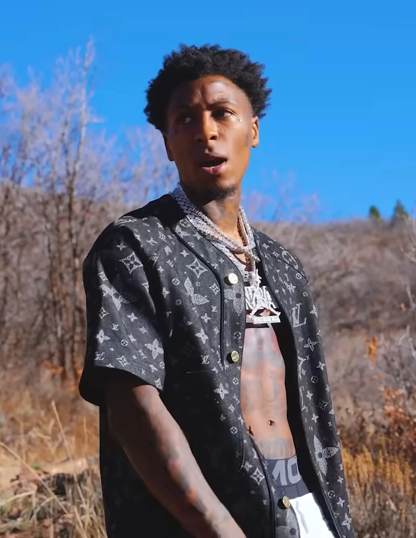 YoungBoy Never Broke Again standing outdoors in a black patterned shirt with layered chains against a blue sky.