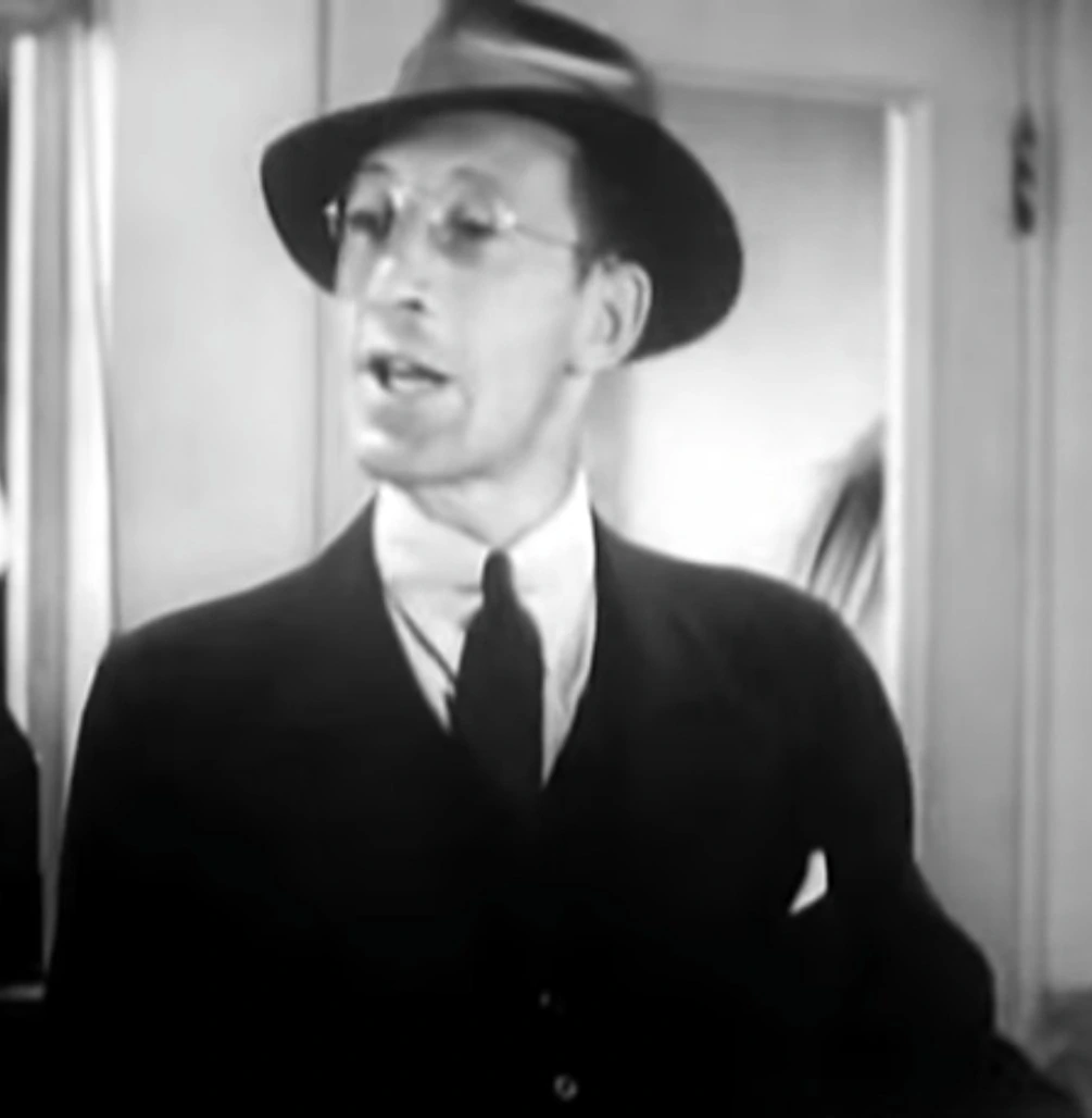 Charles Lane in a black-and-white film still, wearing a suit, tie, hat, and glasses.