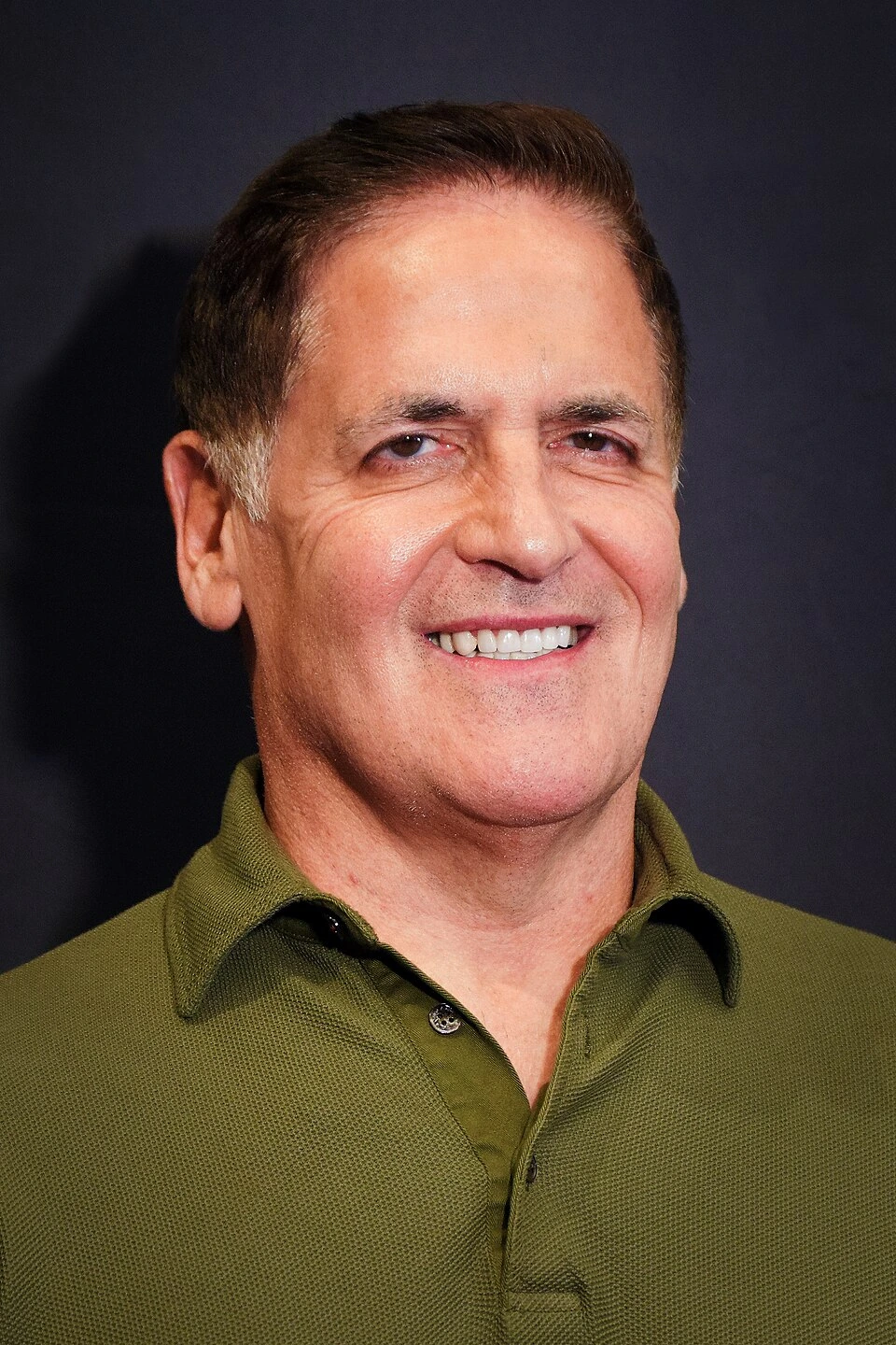 Mark Cuban smiling in a portrait photo, wearing a green shirt against a dark background