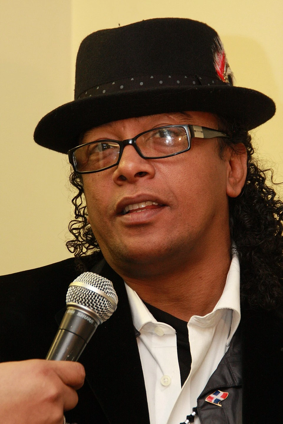 Portrait of Sergio Vargas wearing a black hat and glasses, speaking into a microphone.