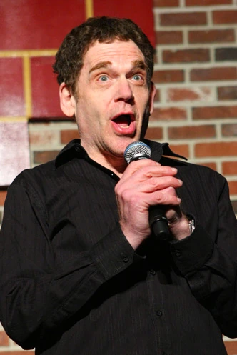 Photo of Charles Fleischer holding a microphone onstage against a brick wall.