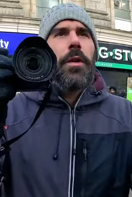 A bearded man in a gray beanie and dark jacket holding a camera outdoors on a city street.