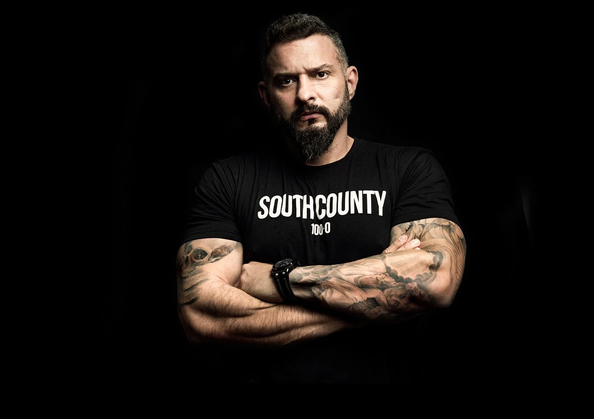 Portrait photo of Andy Frisella standing with arms crossed against a dark background