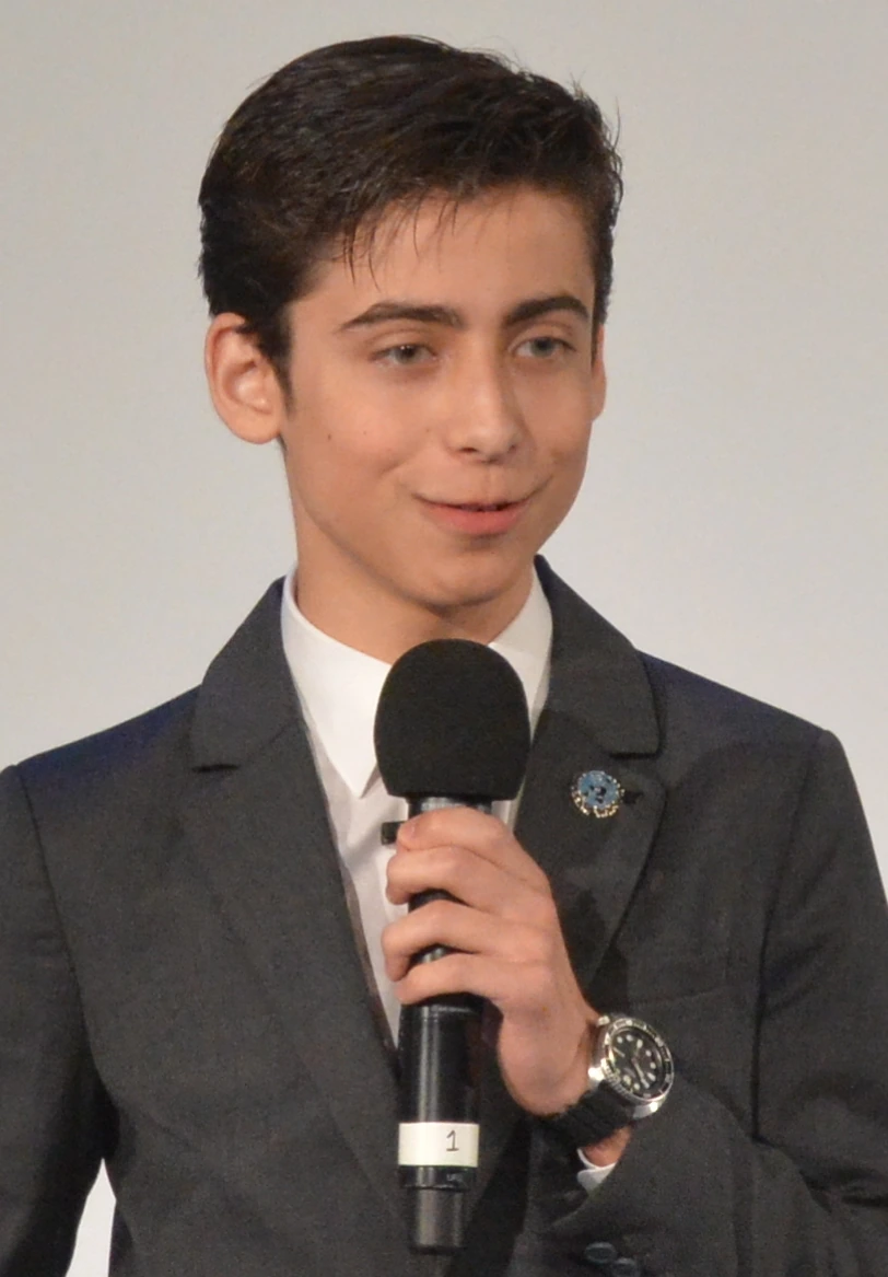 Aidan Gallagher speaking into a microphone in a suit at an event.