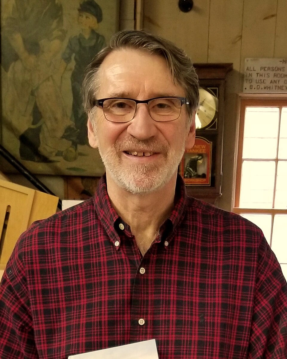 Norm Abram standing indoors in a plaid shirt, smiling at the camera.