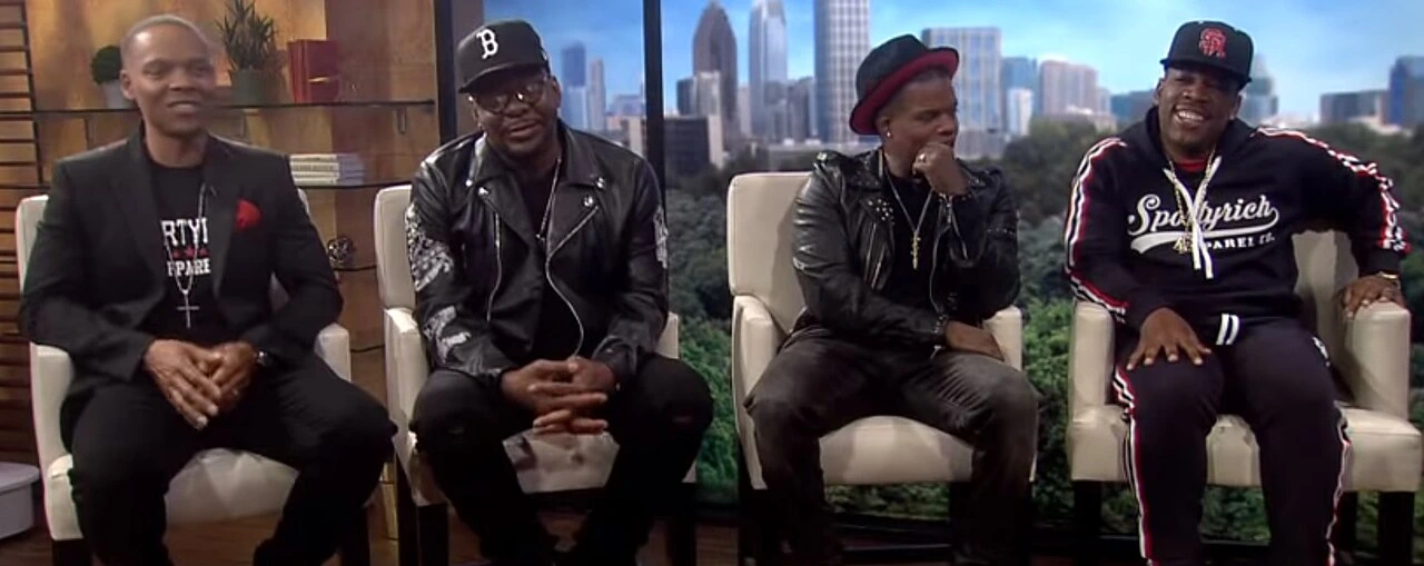Bell Biv DeVoe members seated on a television set interview, with Ricky Bell at far left.