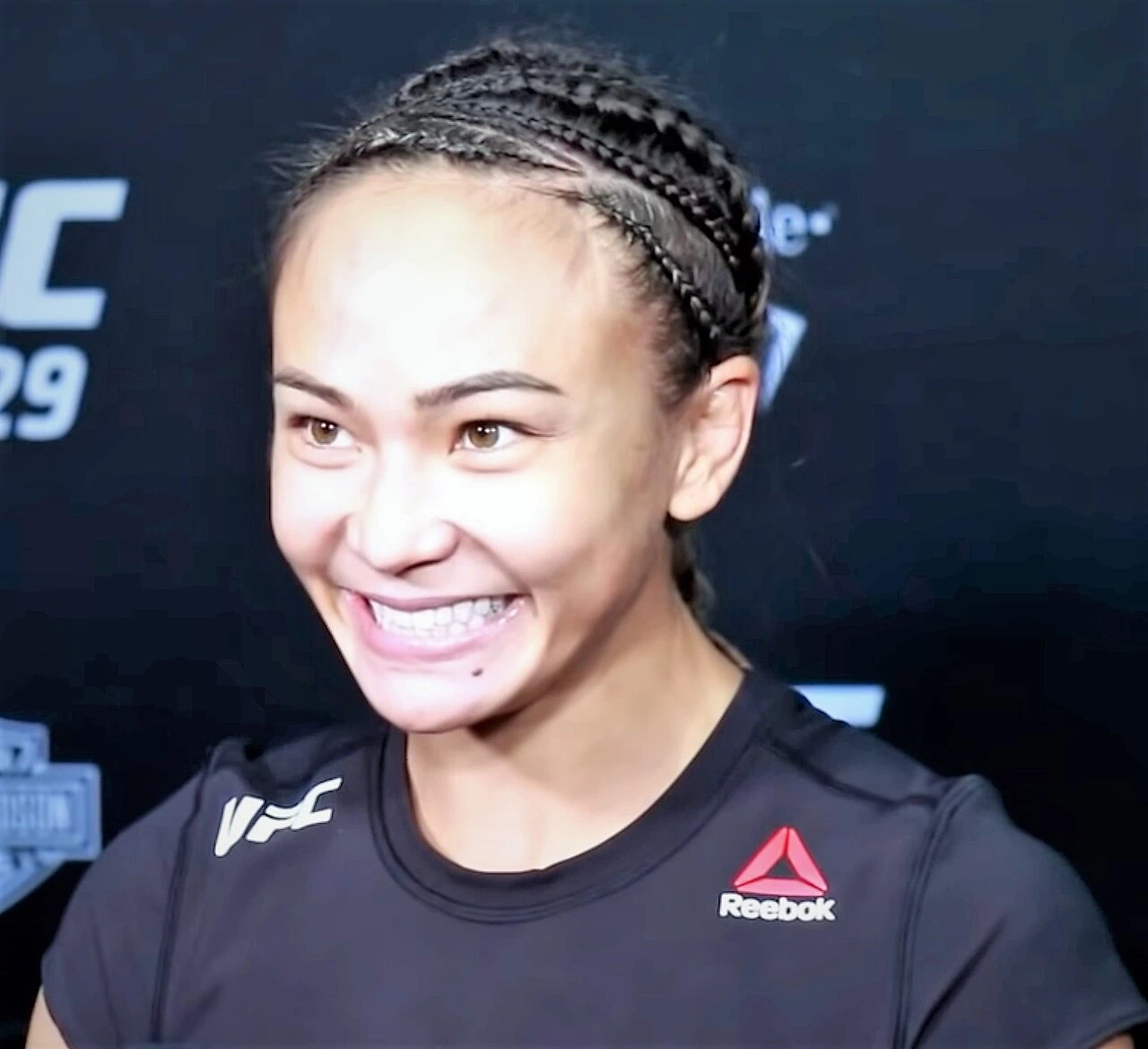 Michelle Waterson smiling at a UFC 229 media event, wearing a black UFC/Reebok shirt and braided hair.