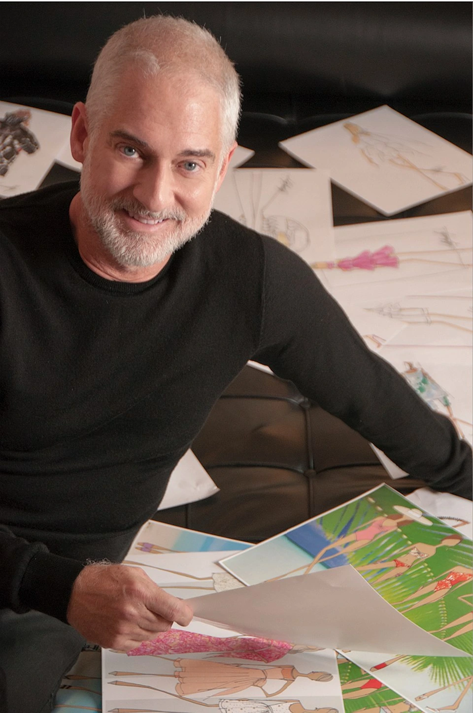 Sully Bonnelly seated indoors among fashion sketches, smiling at the camera