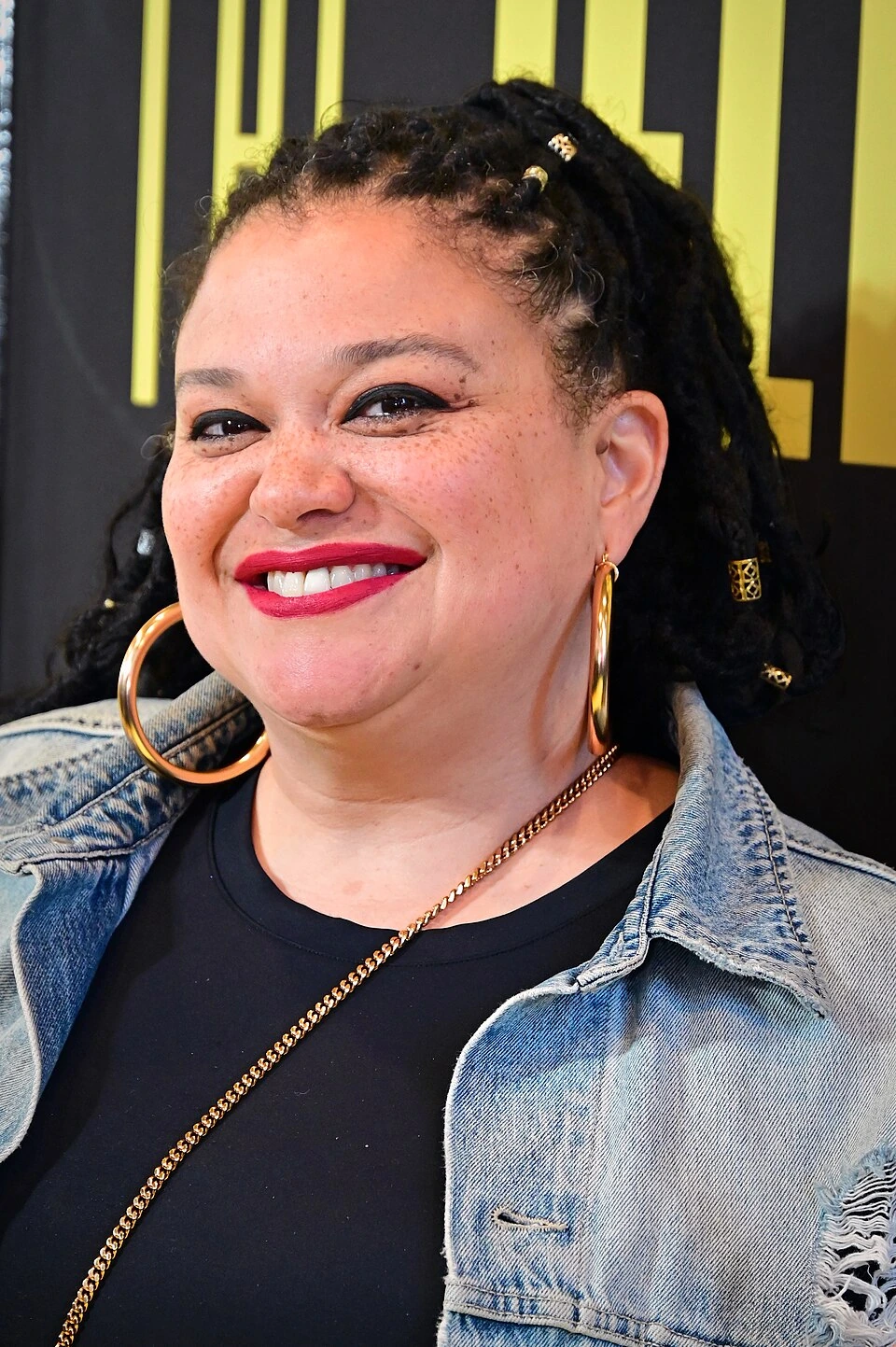 Michelle Buteau smiling at an event, wearing a denim jacket and large hoop earrings.