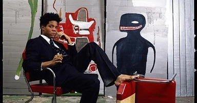 Jean-Michel Basquiat seated in an art studio, holding a cigarette with paintings behind him.