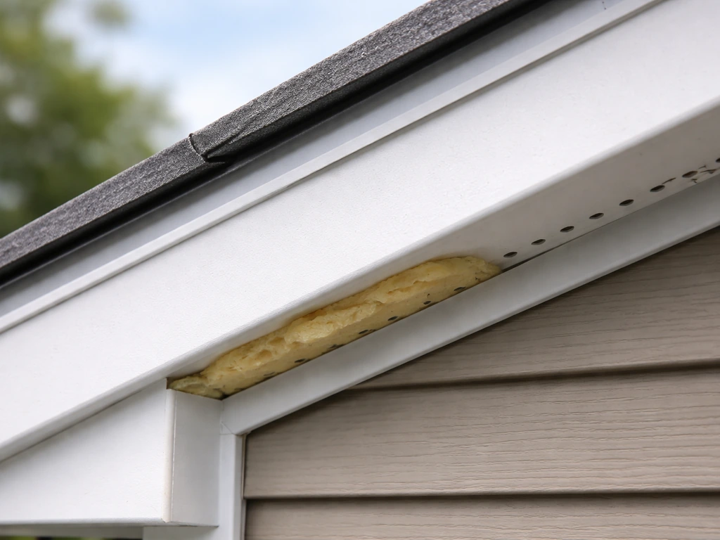 Close-up of a roof overhang weatherproofing seam with caulk, trim, insulation, and small vent/drain openings.