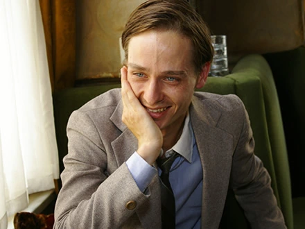 Portrait of Tom Schilling, a German actor, seated indoors and smiling thoughtfully.