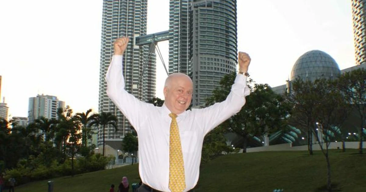 Tom 'Big Al' Schreiter standing outdoors with arms raised in front of city buildings at sunset.