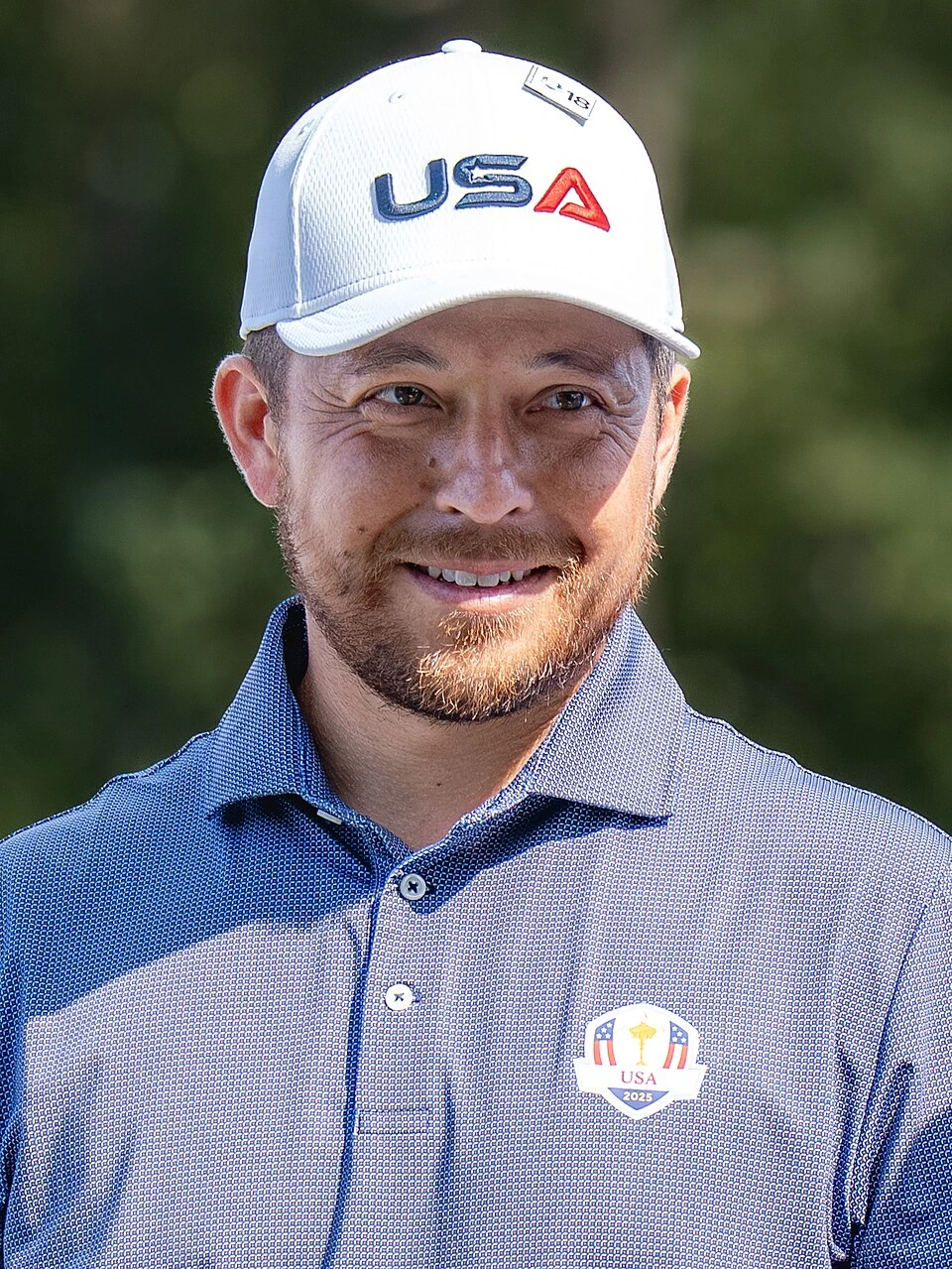 Xander Schauffele at the Ryder Cup in a blue USA team shirt and white cap, smiling in a close-up portrait