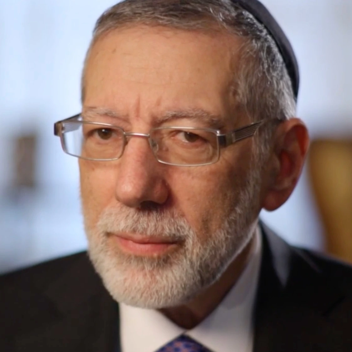 A head-and-shoulders portrait of Avi Schron wearing glasses and a suit, looking toward the camera.
