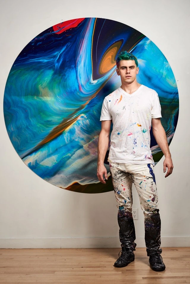Callen Schaub standing in front of a large circular abstract painting in a studio, wearing paint-splattered clothes.