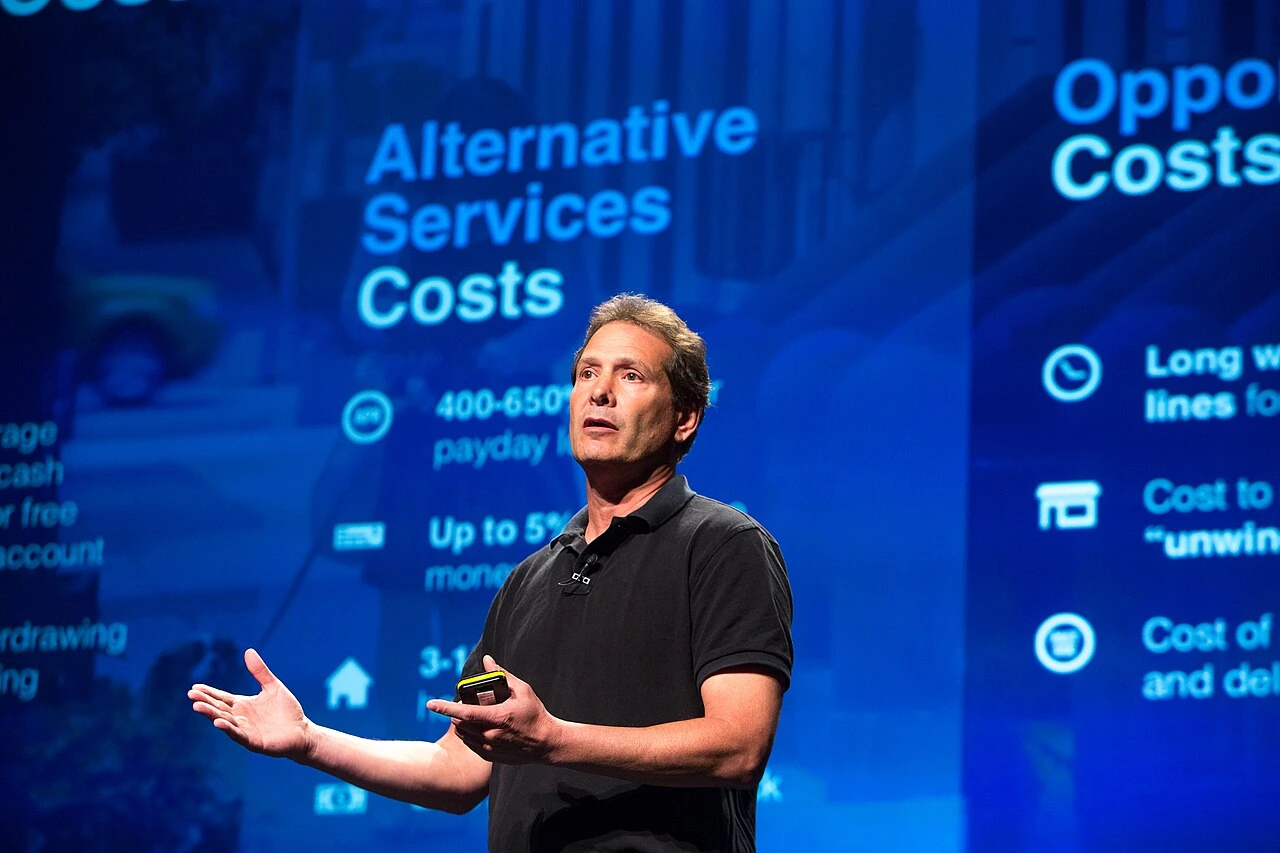 Dan Schulman speaking on stage during a presentation, gesturing with one hand against a blue event backdrop.