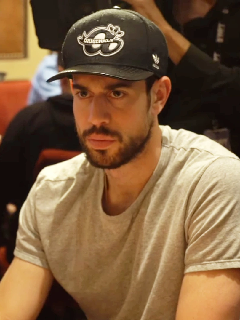 Nick Schulman seated at a poker table wearing a black cap and gray T-shirt
