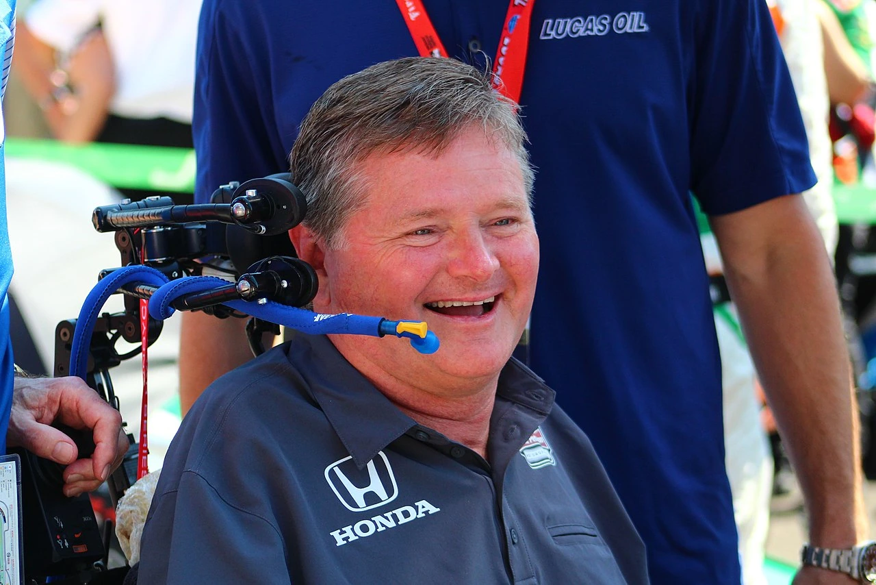 Sam Schmidt smiling in a wheelchair at a racing event, wearing a Honda-branded shirt.