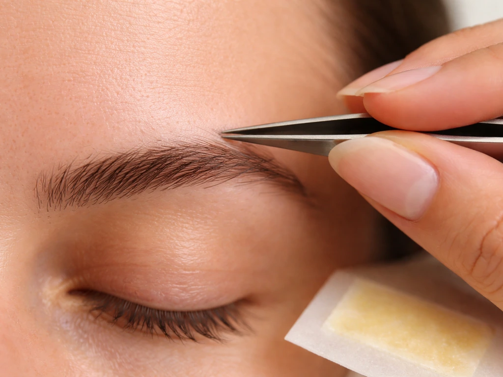 Close-up of tweezers plucking fine hairs from the center of an eyebrow.