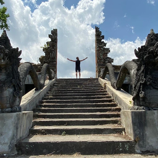 A person standing on stone steps with arms outstretched between ornate carved structures under a cloudy sky.