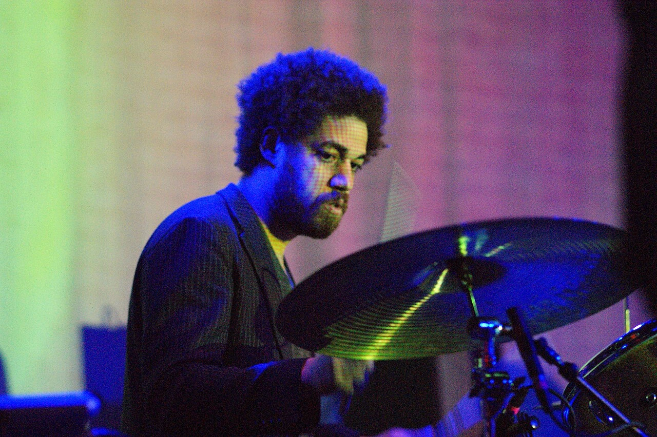 Danger Mouse performing on stage behind a drum kit, lit by blue and purple concert lighting.