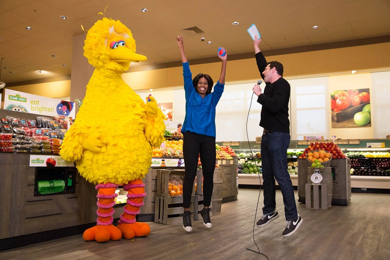 Big Bird standing in a supermarket scene with two people raising their arms nearby