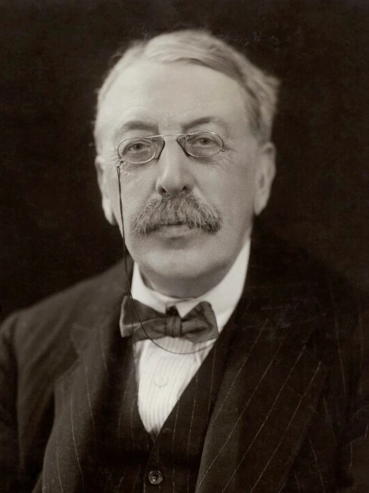 Portrait photograph of composer Charles Villiers Stanford wearing glasses and a bow tie.