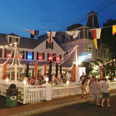 Nighttime exterior view of Patio American Grill & Cocktail Bar with its patio terrace, white fencing, string lights, and
