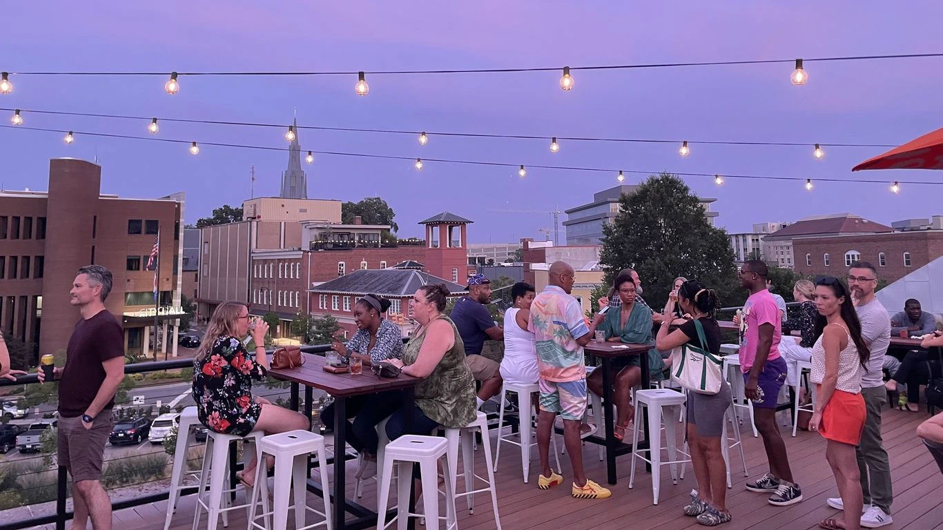 Rooftop patio bar at Unscripted Durham with string lights, guests seated at tables, and the Durham skyline at dusk