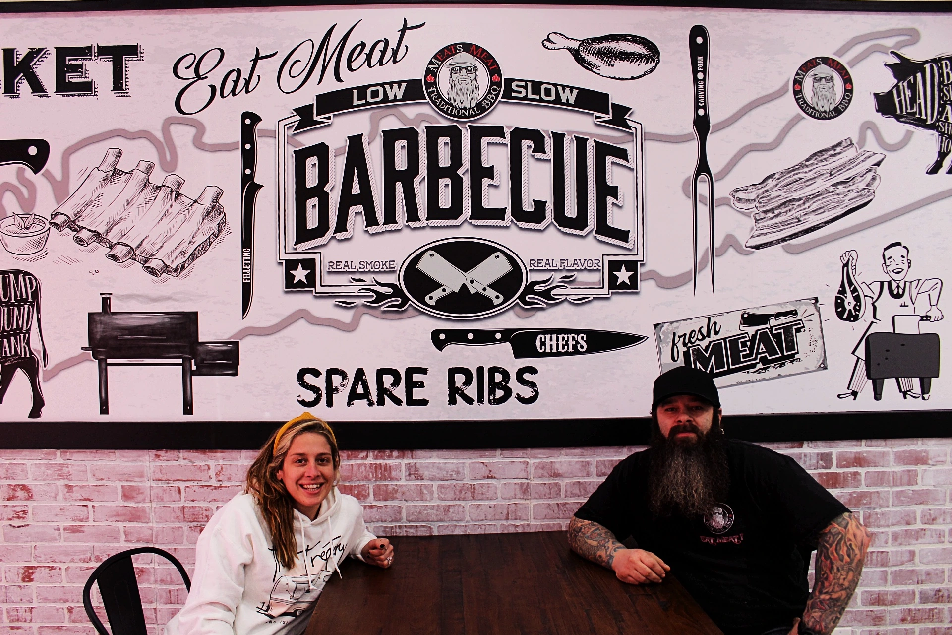 Larry Mondello standing indoors at Meats Meat BBQ in front of a large branded barbecue mural