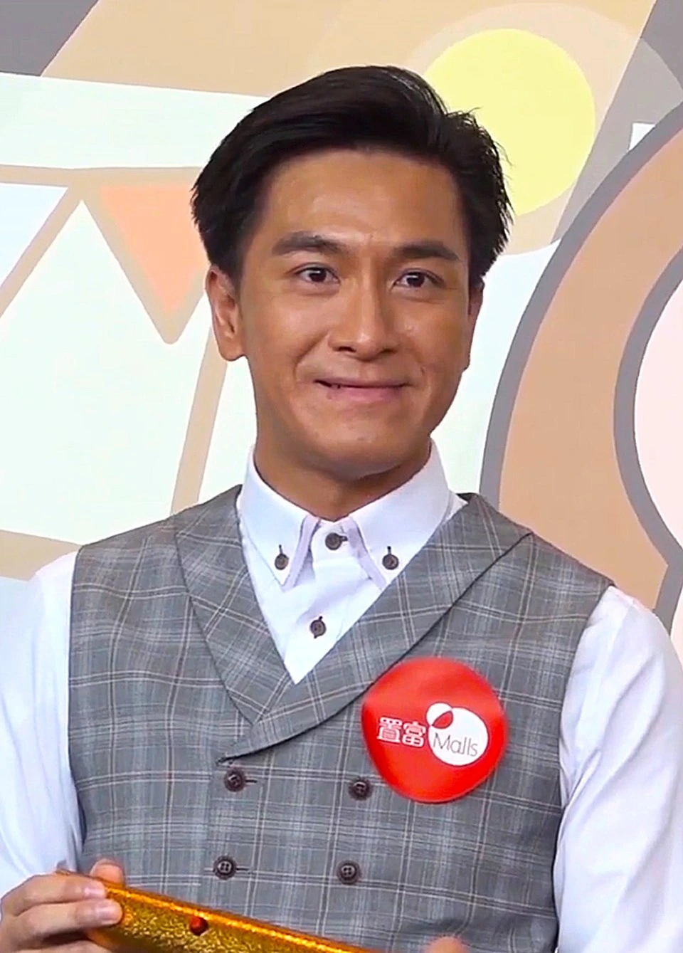 Kenneth Ma Kwok-ming in a studio-style portrait wearing a gray vest and white shirt, standing against a colorful backgro