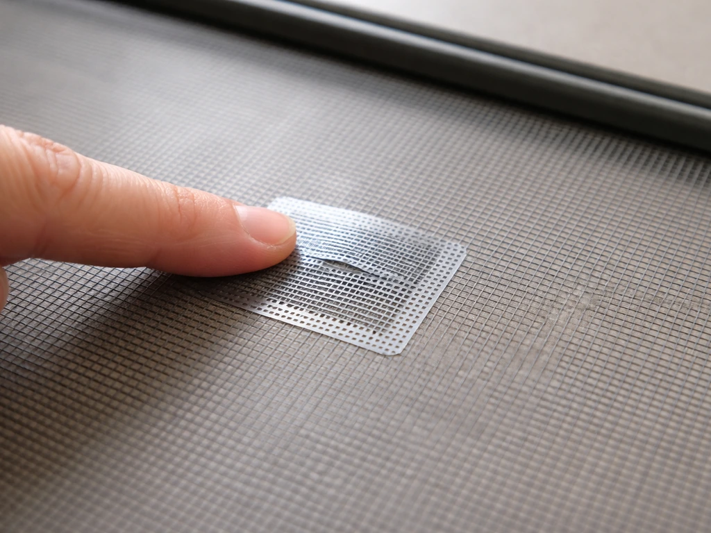 Close-up of a small tear in screen mesh with an adhesive patch pressed over it near the spline/frame.