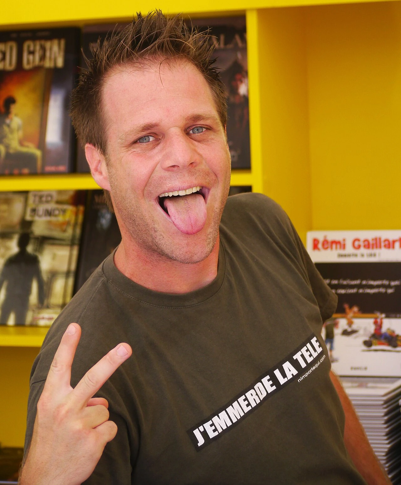 Rémi Gaillard making a playful face and peace sign indoors at an event, with a bright yellow background and posters behi