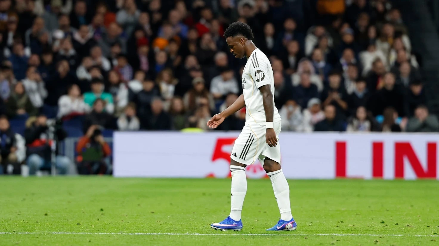 Vinícius Júnior walking on the pitch in a Real Madrid match, in white kit with stadium crowd in the background.