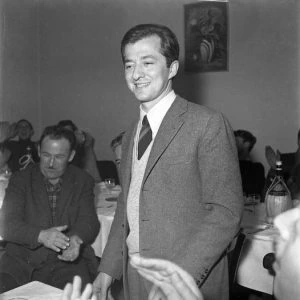 Black-and-white photo of a young Leonardo Del Vecchio standing indoors in a suit and tie, smiling at an event.
