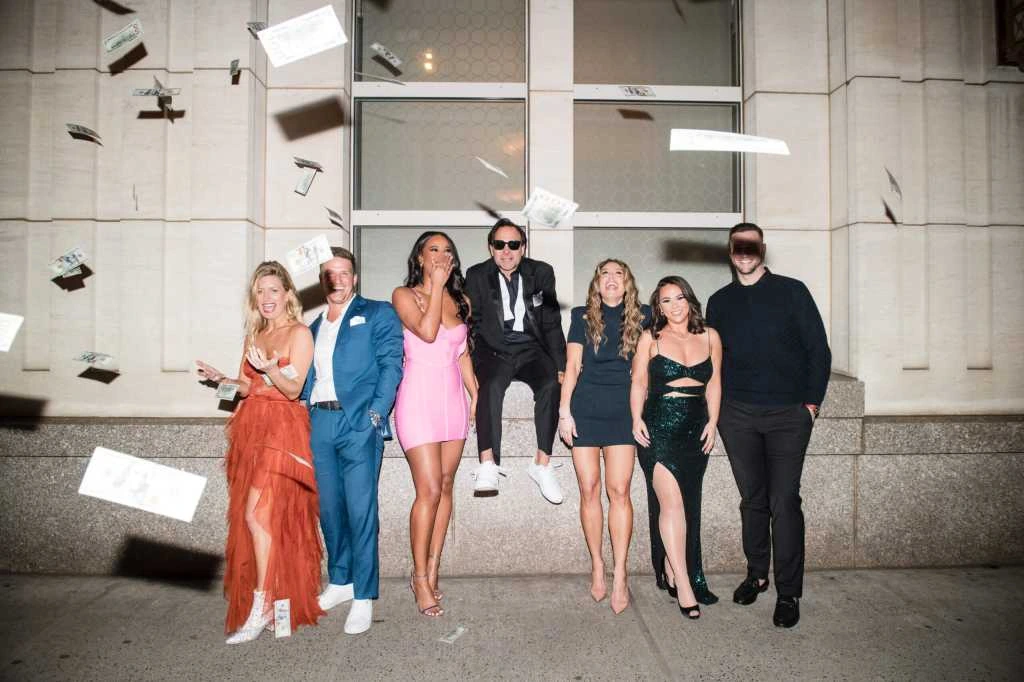 Group photo from a Selling the Hamptons premiere event, showing cast members posing in front of a building as money fall