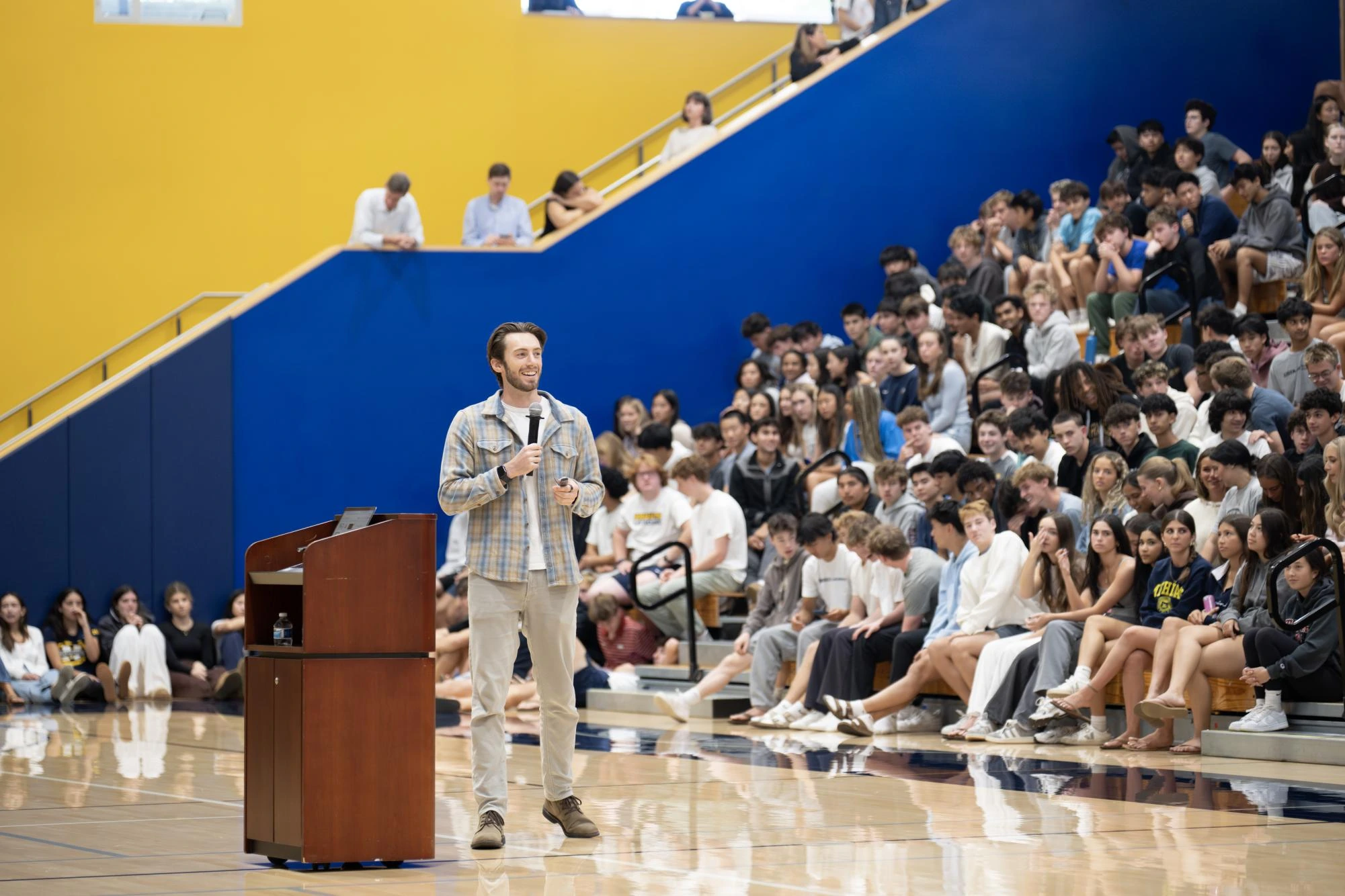 Dino Ambrosi speaking to students in a gymnasium, holding a microphone during a Project Reboot presentation.