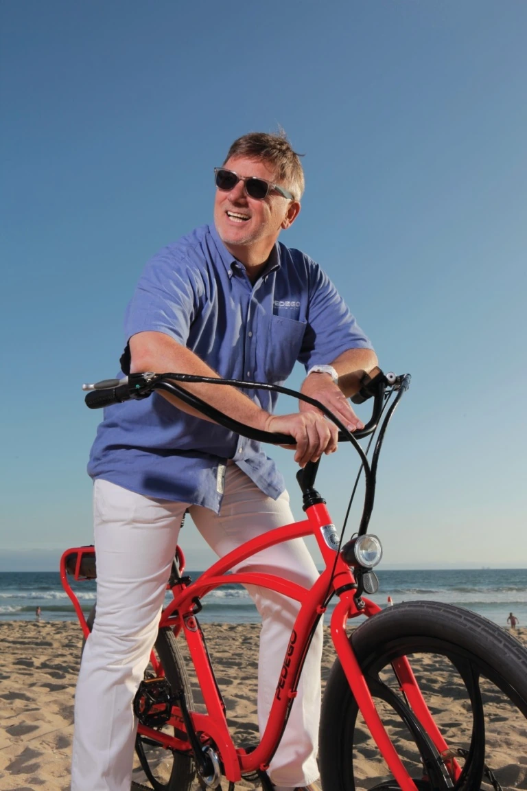 Don DiCostanzo standing with a red Pedego electric bike at the beach