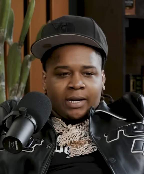 YTB Fatt speaking into a microphone during an interview, wearing a cap and chain.