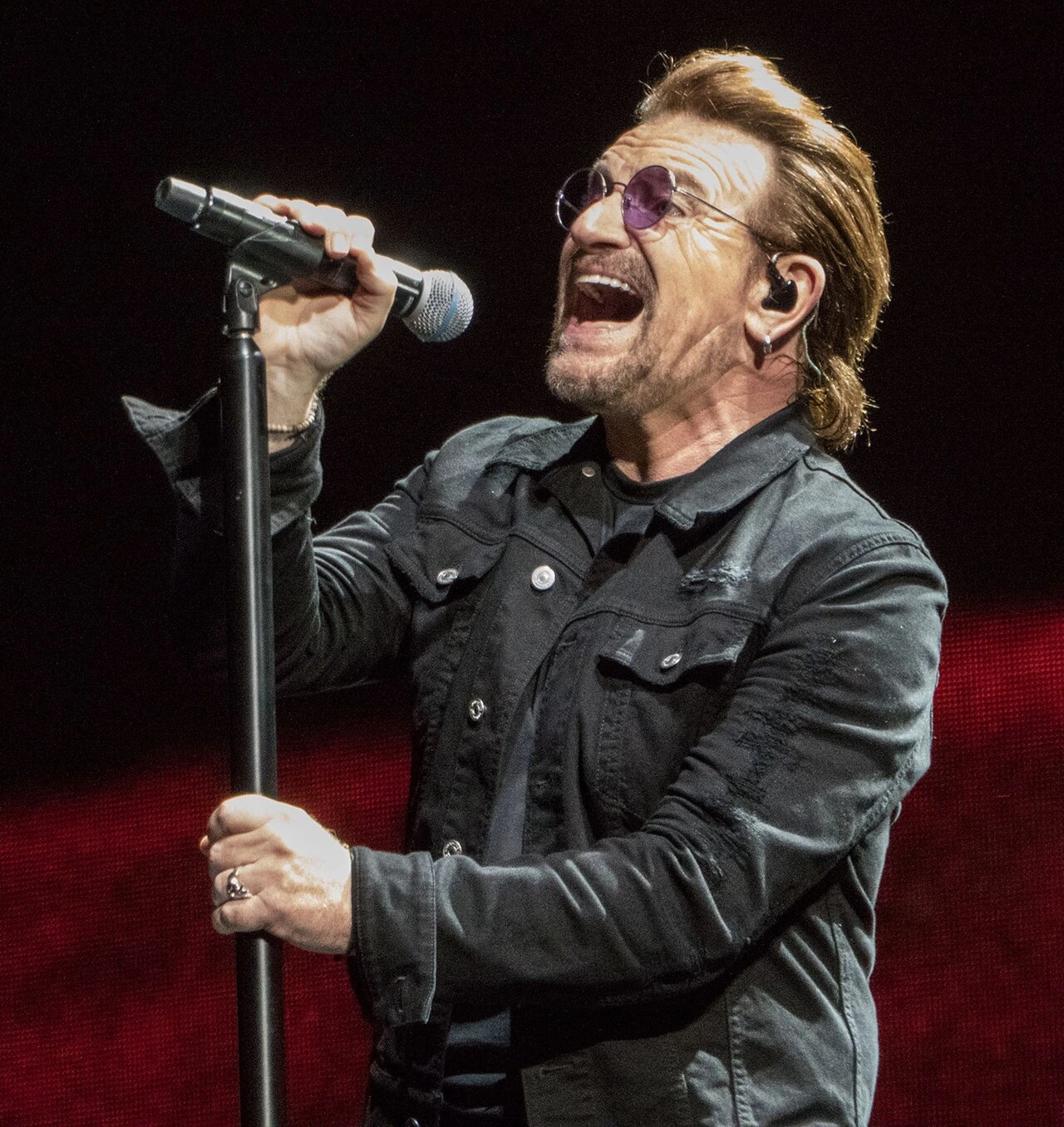 Bono singing on stage during the Joshua Tree Tour 2017, holding a microphone and wearing tinted glasses.