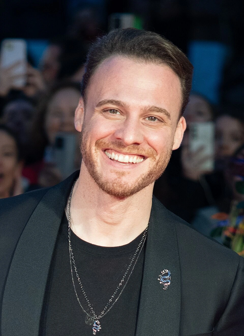Kerem Bürsin smiling at a red carpet event, wearing a black jacket and shirt.