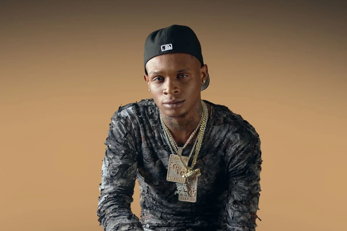 Portrait of Los Angeles rapper HoodTrophy Bino wearing a black cap and layered chains against a plain background