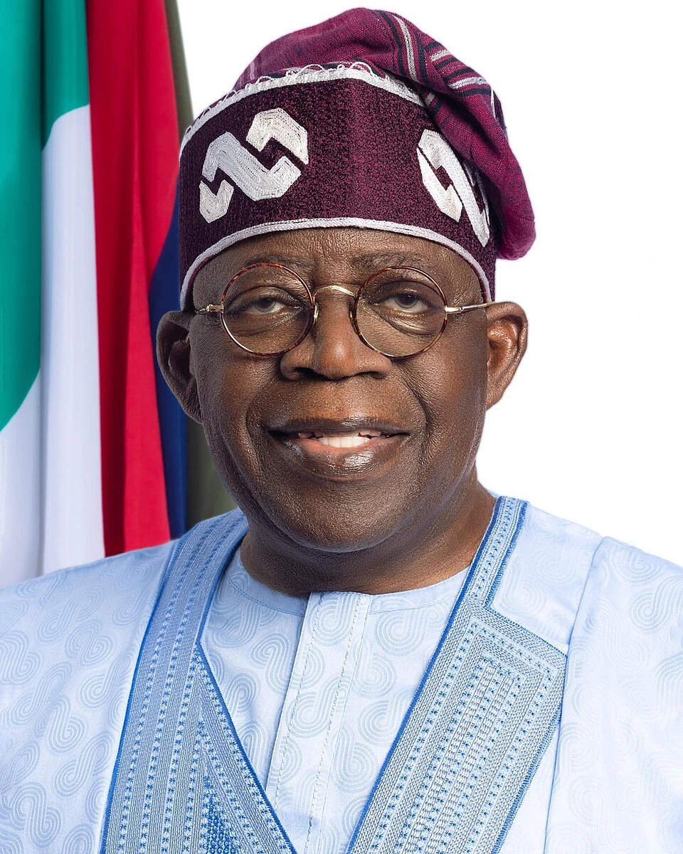 Portrait photo of Bola Tinubu wearing traditional attire and glasses, with the Nigerian flag in the background.