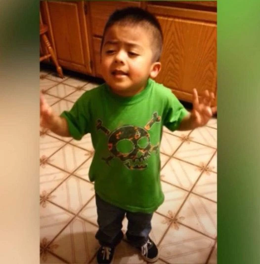 A young boy in a green shirt standing indoors on a tiled floor, from the Listen Linda viral video.