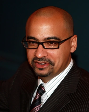 Portrait photo of Junot Díaz in a suit and glasses against a dark background