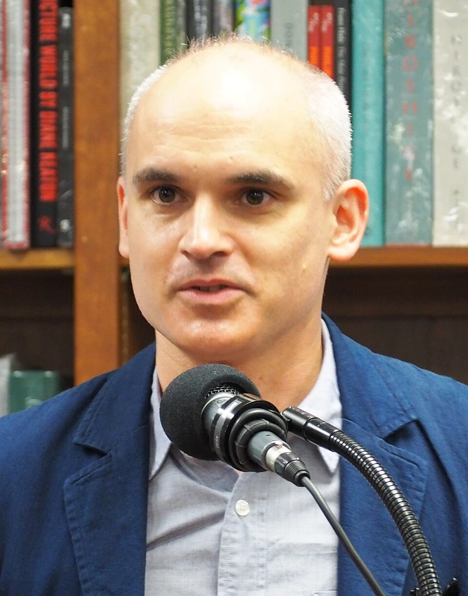 Hernán Díaz speaking at a microphone in front of bookshelves